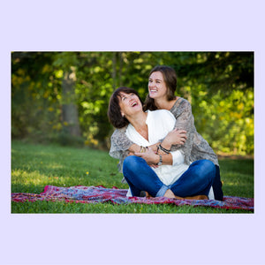 Mother and daughter sitting on a blanket in a park, hugging and smiling.