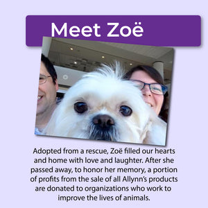 White dog being held by two people with a purple banner above that reads, "Meet Zoë" and text below that reads, "Adopted from a rescue, Zoë filled our hearts and home with love and laughter. After she passed away, to honor her memory, a portion of profits from the sale of all Allynn's products are donated to organizations who work to improve the lives of animals."